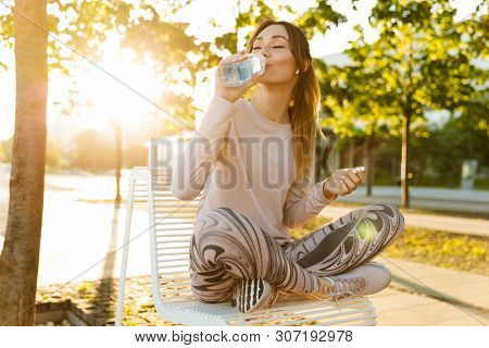 Cheerful young fitness woman relaxing outdoos, sitting on a bench, drinking water