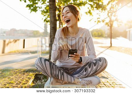 Cheerful excited young fitness woman relaxing outdoos, sitting on a bench, using mobile phone