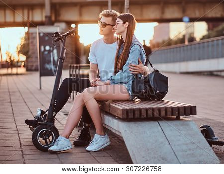 Young Beautiful Romantic Couple With Electric Scooters Is Sitting On The Bench Near Brige At Sunset 