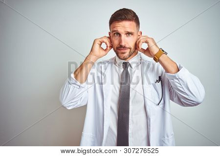Young handsome doctor man wearing white profressional coat over isolated background covering ears with fingers with annoyed expression for the noise of loud music. Deaf concept.
