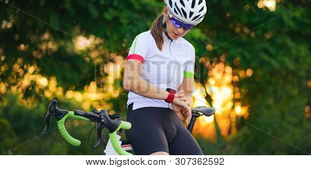 Young Woman Road Cyclist Resting with Bicycle and Using Multisport Smartwatch at Sunset. Closeup of Hands with Fitness Tracker. Training and Sports Concept.