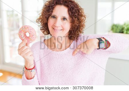 Senior woman eating pink sugar donut with surprise face pointing finger to himself
