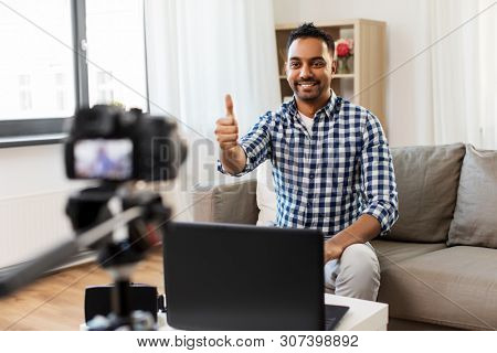 blogging, videoblog and people concept - indian male blogger with camera recording video blog and showing thumbs up at home