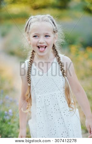 Funny Girl Holding Hair Plaits. Small Cute Girl With Long Blonde Hair Showing Tongue Outdoor. Kid Ex
