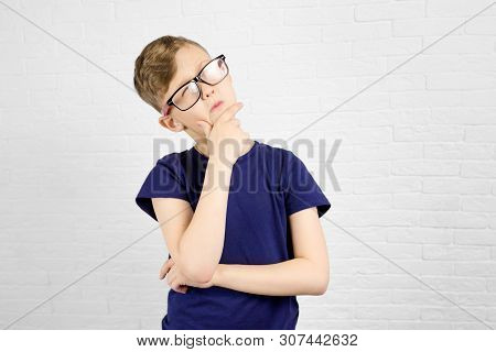 Thoughtful Schoolboy Wearing Eyeglasses Standing Against White Brick Wall. Back To School Concept.