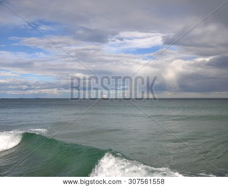 A Clear Blue Sky Is Visible Through The Gray Clouds On The Sea. In The Foreground A Green Wave. In T