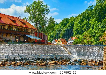 Cesky Krumlov (Czech Krumlov), Czech Republic. Antique town on river Vltava. Picturesque landscape. Cascade dam with stones. Colourful houses on bank among green trees. Sunny summer day with blue sky.
