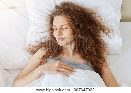 Indoor Shot Of Young Female With Curly Hair, Laying In White Bed With Closed Eyes And Happy Facial E