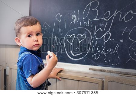 Little Boy Standing Near School Blackboard With A Piece Of Chalk