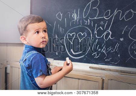 Little Boy Standing Near School Blackboard With A Piece Of Chalk