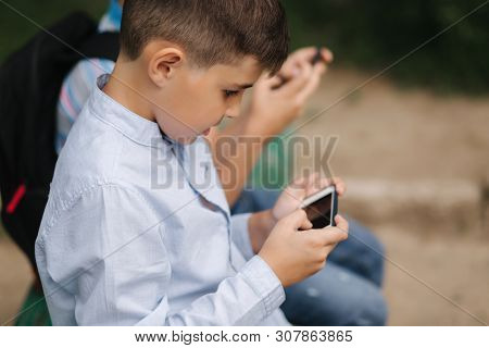 Two Boy Sitting On The Bench And Play Online Games. One Boys With Backpack. Young Boys Use Their Pho