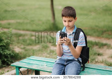 Young Boy With Backpack Sitting On The Bench And Play Online Games During School Break