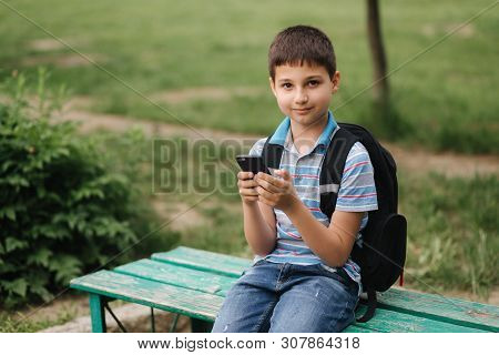 Young Boy With Backpack Sitting On The Bench And Play Online Games During School Break