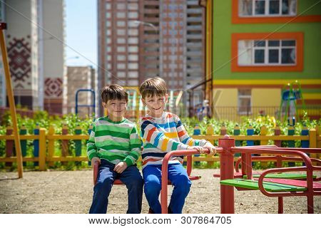 Two Cheerful Boys In T-shirts Play On A Big Iron Carousel On The Playground At The Summer Or Spring