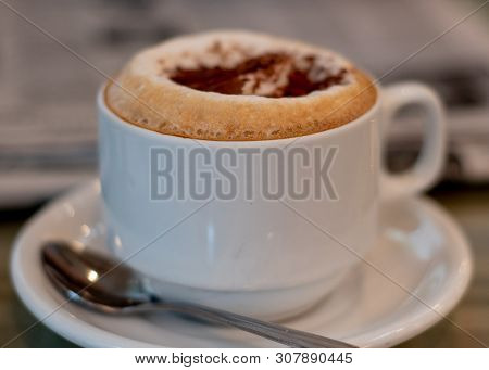 Selective Focus On Cappuccino On Table At A Cafe Filling The Frame, With Newspapers In The Backgroun