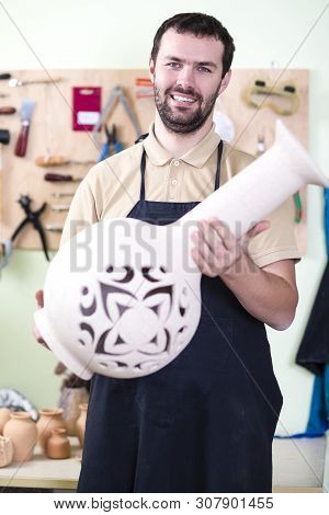 Happy Cacucasian Male Potter In Workshop Posing With Big Clay Jar Against Instruments Board. Vertica