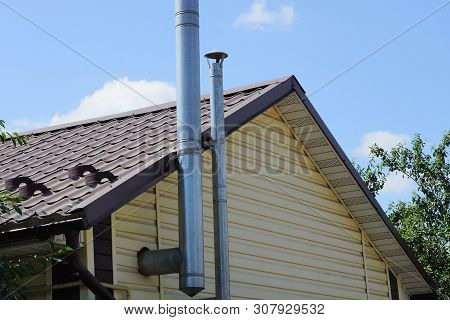 Two Metal Chimney Pipes In The Brown Attic Of The House Against The Sky
