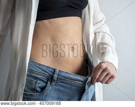 A Slender Woman With A Bare Stomach Shows How She Lost Weight, Holding Her Jeans With Her Hand. The 