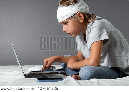 Little Caucasian Blond Boy With A Head Injury And Bandage Is Sitting On The Bed And Using Laptop. Re