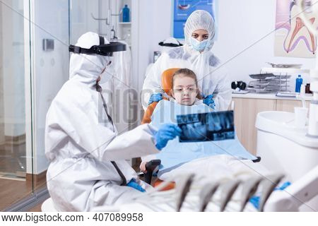 Dentist With Face Shield And Ppe Suit In Dental Clinic Discussing About Caries Using Radiography. St