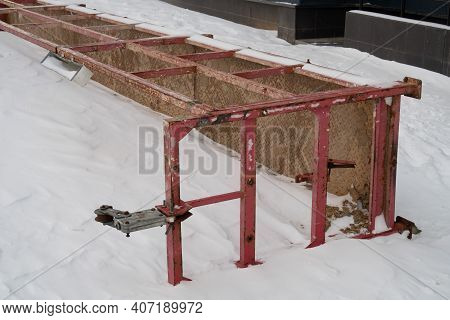 Red Rusty Fromt Construction Cradle Lies On Its Side In The Snow Next To The Foundation Of The House