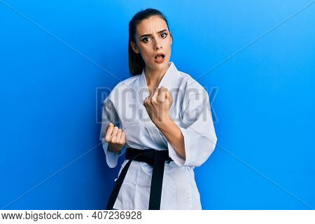 Beautiful brunette young woman wearing karate fighter uniform with black belt doing attack pose in shock face, looking skeptical and sarcastic, surprised with open mouth 