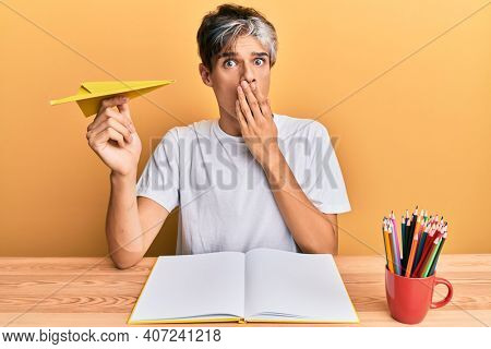 Young hispanic man studying and holding paper airplane sitting on the table covering mouth with hand, shocked and afraid for mistake. surprised expression 