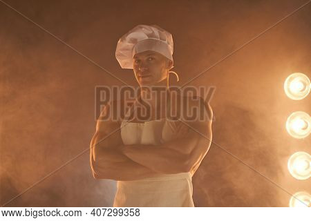 Portrait Of Muscular Chef Wearing White Apron And Chef Hat, Posing With Folded Arms On Smoky Backgro