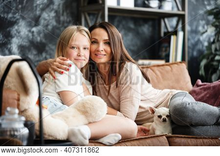 Mother and daughter relationship concept. A woman and a teenage girl are sitting in front of the TV on the couch and happy embarcing. The family spends time at home. Little dog lying on laps.