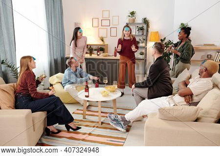 Group of young cheerful intercultural friends in smart casualwear gathered around small white round table in living-room and playing name game
