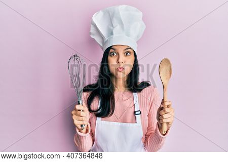 Young hispanic woman wearing baker uniform holding spoon and whisk puffing cheeks with funny face. mouth inflated with air, catching air. 