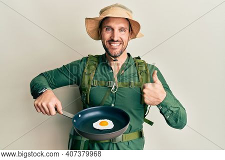 Handsome man with beard wearing explorer hat cooking fried egg smiling happy and positive, thumb up doing excellent and approval sign 