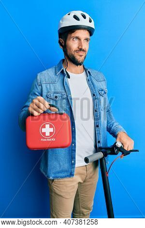Young man standing on scooter wearing helmet holding first aid kit smiling looking to the side and staring away thinking. 