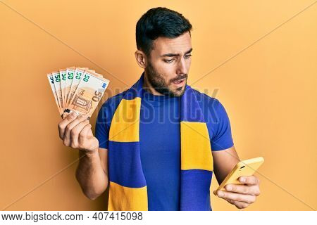 Young hispanic man football supporter using smartphone holding 50 euro banknotes in shock face, looking skeptical and sarcastic, surprised with open mouth 