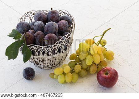 Just Picked Ripe Plums In Wicker Basket And A Bunch Of Ripe White Grapes, An Apple On The White Stru
