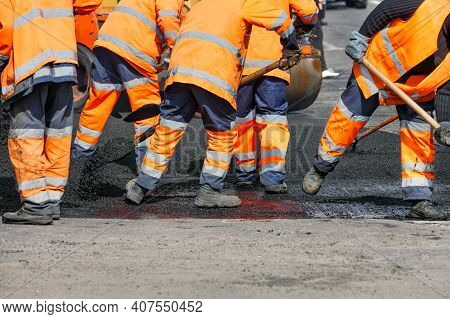 A Team Of Road Workers In Orange Reflective Uniforms Use Shovels To Scatter Some Of The Asphalt To R