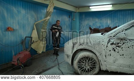 A Caucasian Man Works In A Car Wash Service. A Man In A Workers Uniform Sprays A Car With Detergent
