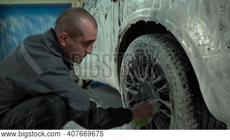 A Caucasian Man Works In A Car Wash Service. A Man In A Workers Uniform Washes The Wheel Of A Car Th