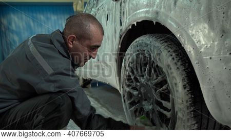 A Caucasian Man Works In A Car Wash Service. A Man In A Workers Uniform Washes The Wheel Of A Car Th