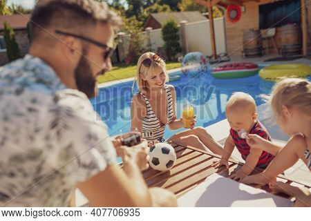 Beauitful Young Family Spending Hot Sunny Summer Day By The Swimming Pool, Parents Relaxing And Drin