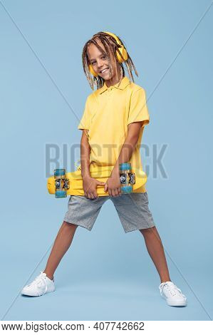 Confident Little Boy With African Dreads With Headphone And Skateboard Posing Over Blue Background.