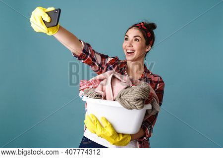 Joyful caucasian woman with dirty clothes taking selfie on cellphone isolated over blue background