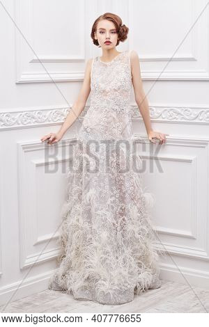 Wedding fashion. Full length portrait of a beautiful young woman in a long evening white dress posing in a classic white interior. Evening makeup and hairstyle. 
