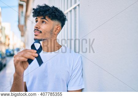 Young arab man with serious expression holding mouring black ribbon leaning on the wall.