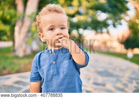 Sad little boy putting fingers on mouth touching gums because toothache at the park on a sunny day. Beautiful blonde hair male toddler in pain for new baby teeth outdoors