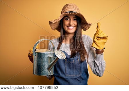 Young beautiful brunette farmer woman wearing apron and hat holding watering can surprised with an idea or question pointing finger with happy face, number one