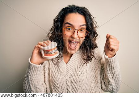 Young beautiful woman with curly hair holding plastic teeth with dental baces screaming proud and celebrating victory and success very excited, cheering emotion