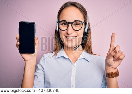 Beautiful call center agent woman working using headset holding smartphone holding screen surprised with an idea or question pointing finger with happy face, number one