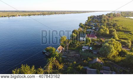 Drone Fly Over Waving River Of Blue Color Surrounded By Local Village With Various Buildings And Wet