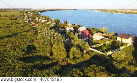 Drone Fly Over Waving River Of Blue Color Surrounded By Local Village With Various Buildings And Wet